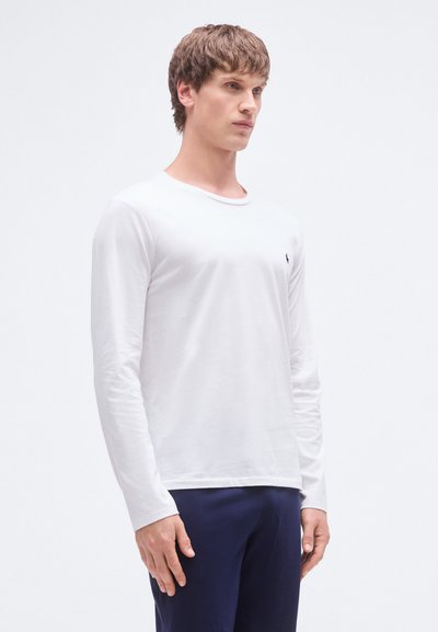 Hvit skjorte med lange ermer, rund hals, myk tekstur og en liten svart logo på venstre bryst, kombinert med marineblå bukser.