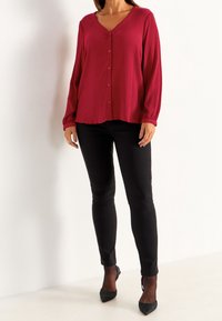 Blusa cremisi con bottoni frontali, scollatura a V e maniche lunghe, abbinata a pantaloni neri aderenti e tacchi neri su uno sfondo neutro.