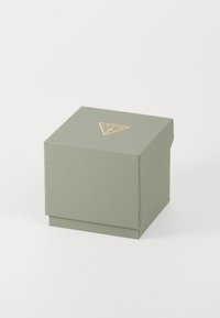 Lille tekstureret grå-grøn firkantet boks med en gylden Guess-logo-triangel på toppen, lukket og mod en hvid baggrund.