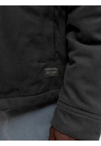 Gros plan d'une personne portant une veste noire Jack & Jones avec des coutures visibles et une petite étiquette de marque près de l'ourlet.