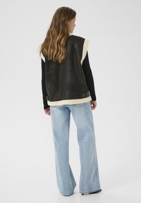 Žena s dugom valovitom kosom nosi crnu majicu dugih rukava, tamni krzneni prsluk i svijetloplave široke traperice dok stoji naslonjena na bijelu pozadinu.