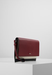 Rektangulær tri-farvet taske med bordeaux flap, sort bund, beigefarvet midtersektion og en matchende bordeaux håndledsrem på en hvid overflade.