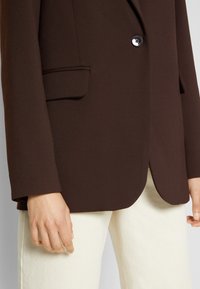 Blazer marrone con due tasche frontali e chiusura a un bottone, realizzato in tessuto testurizzato. Abbinato a pantaloni avorio.