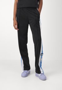 Svarta träningbyxor med vita och blå ränder på sidorna, elastisk midja och en varumärkespatch. Matchas med ljuslila sneakers.