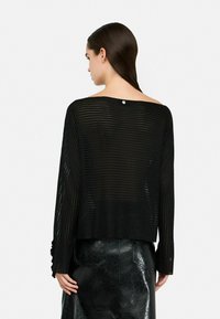 Črno prugasta pletena majica z off-shoulder obliko, dolgimi rokavi in vidnim grbom na hrbtu, v kombinaciji z sijočo črno krilom.