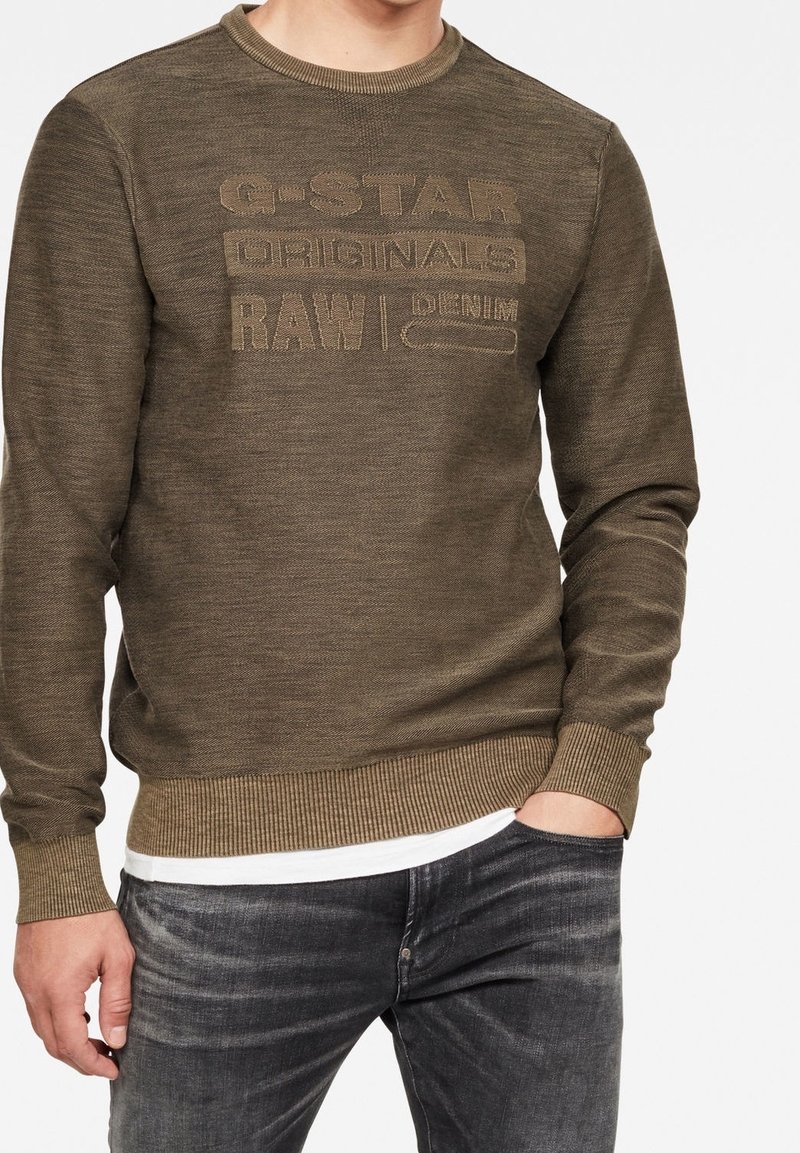 Man med en olivgrön stickad tröja med texten "G-STAR ORIGINALS RAW DENIM", i kombination med blekta svarta jeans och en vit undertröja.