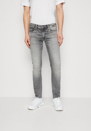 Jeans grigi slim-fit con una finitura sbiadita, cinque tasche, passanti per cintura e chiusura con bottone, abbinati a scarpe da ginnastica bianche e un orologio da polso nero.