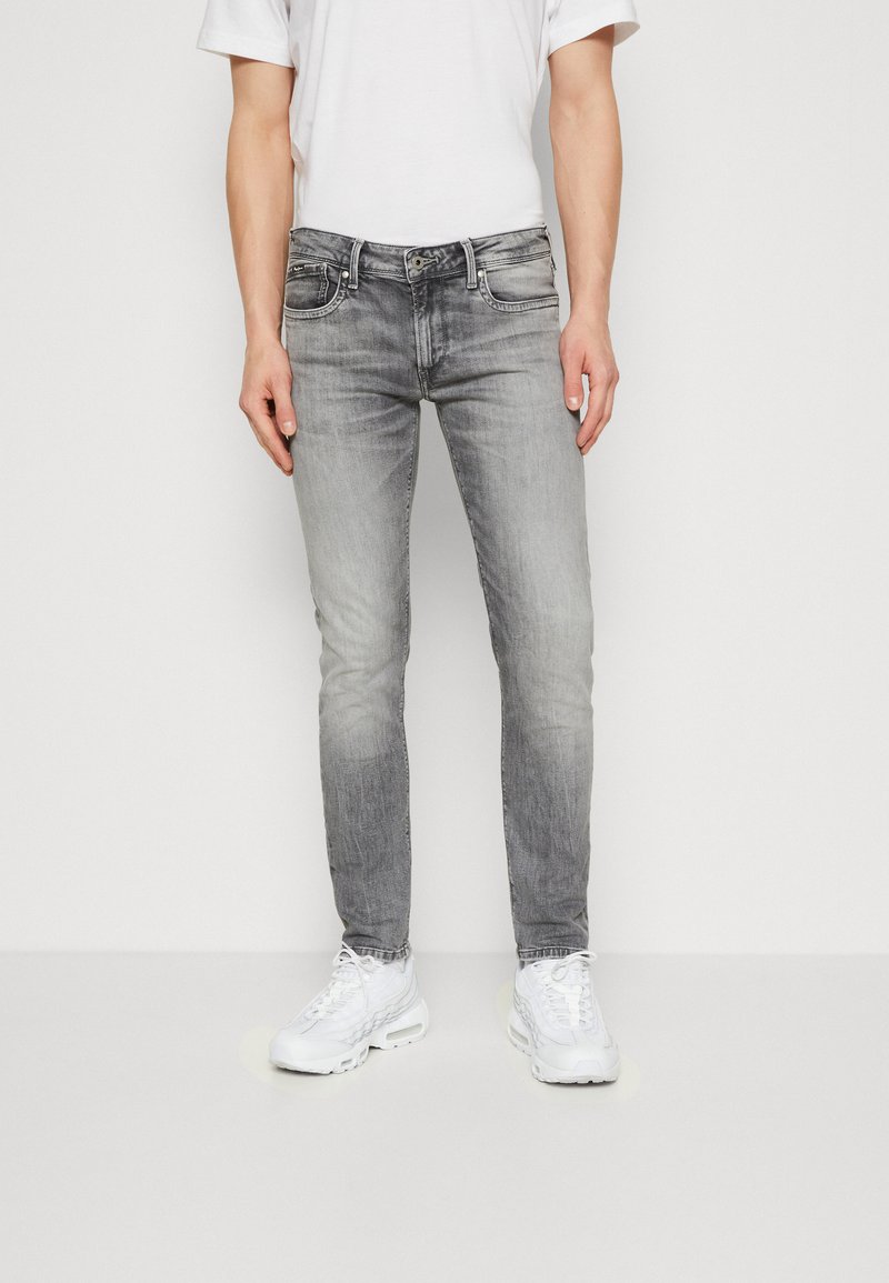 Jeans ajustados grises con un acabado deslavado, cinco bolsillos, trabillas para cinturón y cierre de botones, combinados con zapatillas deportivas blancas y un reloj de pulsera negro.