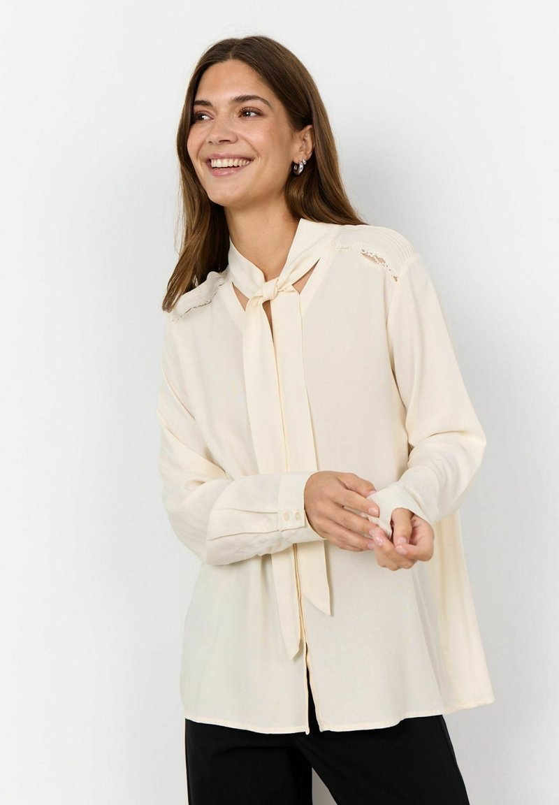 Blusa color crema hecha de una tela ligera, con un escote atado, puños con botón y detalles de encaje en los hombros.