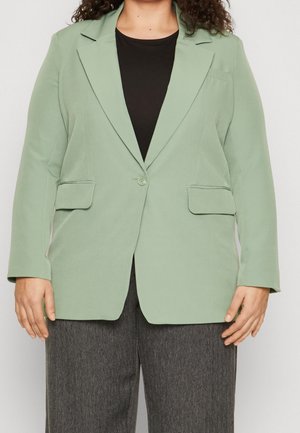 Persoon draagt een lichtgroene blazer met één knoop over een zwart topje en grijze, gestructureerde broek, staand tegen een effen achtergrond.