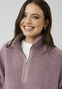 Pull en polaire de couleur violet clair, tissu texturé, design à demi-zip et col. Porté sur une chemise blanche, avec des accents de couture simples.