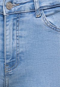 Šviesiai mėlyni džinsai artimas planas, rodantis mygtuką, užtrauktuką, diržo kilpelę ir priekinę kišenę su matomomis siuvimo detalėmis.