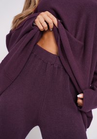 Lila Strickpullover und passende Hose, mit lockerer Passform und weichem Material. Der Pullover ist nach oben gezogen, um die Haut an der Seite zu zeigen.