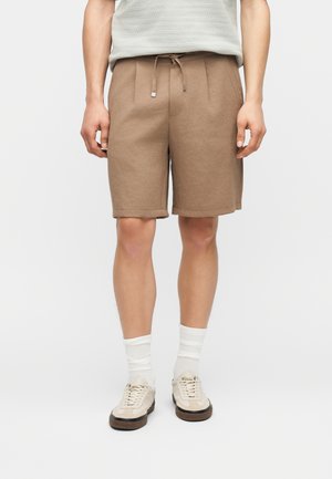 Άνδρας που φοράει ανοιχτό καφέ σορτς με κορδόνια, λευκές κάλτσες και μπεζ αθλητικά παπούτσια, στέκεται μπροστά σε έναν απλό ανοιχτό φόντο.