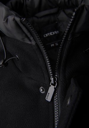 Gros plan sur la fermeture éclair d'une veste d'hiver noire partiellement ouverte, avec la marque "ombre" sur la tirette de la fermeture éclair et l'étiquette intérieure.