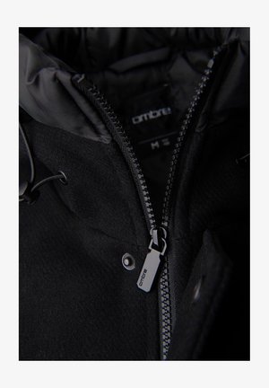 Primo piano di una zip di un giubbotto invernale nero parzialmente aperta, con il marchio "ombre" sul cursore della zip e sull'etichetta interna.