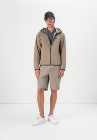 Beige ritsjack met zwarte accenten, gecombineerd met lichtbeige shorts en witte sneakers. Het model draagt ook een zwarte pet.