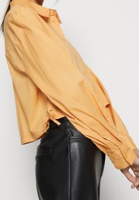 Chemise courte à manches longues en coton orange clair, avec des boutons sur le devant et un détail de noeud à l'ourlet ; associée à un pantalon en simili cuir noir.