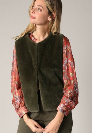 Olijfgroene nepbont vest met een hoge kraag en zonder sluiting aan de voorkant, gelaagd over een bloemenpatroon, rode blouse met lange mouwen.