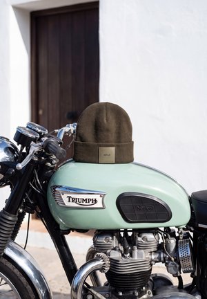 Un bonnet marron Replay repose sur le réservoir vert menthe d’une moto Triumph classique garée à côté d’une porte.