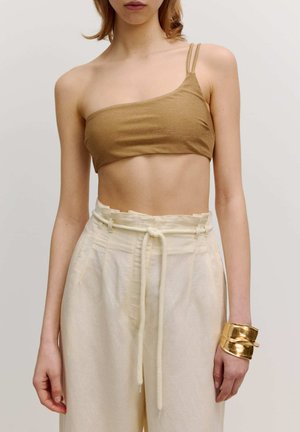 Bronzefarbenes metallisches Crop-Top mit dünnen Trägern, kombiniert mit hellbeigen, schmal zulaufenden Hosen und einem goldenen Armreif. Glatte Textur, minimalistisches Design.
