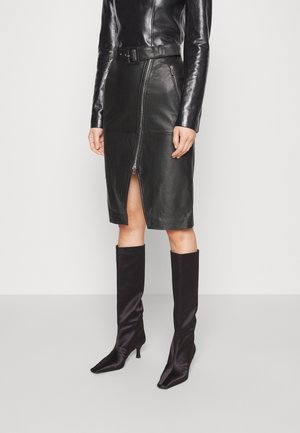 Musta nahkainen midi-hame, jossa on etuvetoketju, sivutaskut ja vyötäröllä vyö. Yhdistetty mustiin polven korkeisiin satiinisiin piikkikorkokengillä.