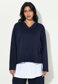 Camiseta de manga larga azul marino con escote en V, de corte relajado y apariencia de capas sobre una camisa blanca, con tejido suave y una ligera textura.
