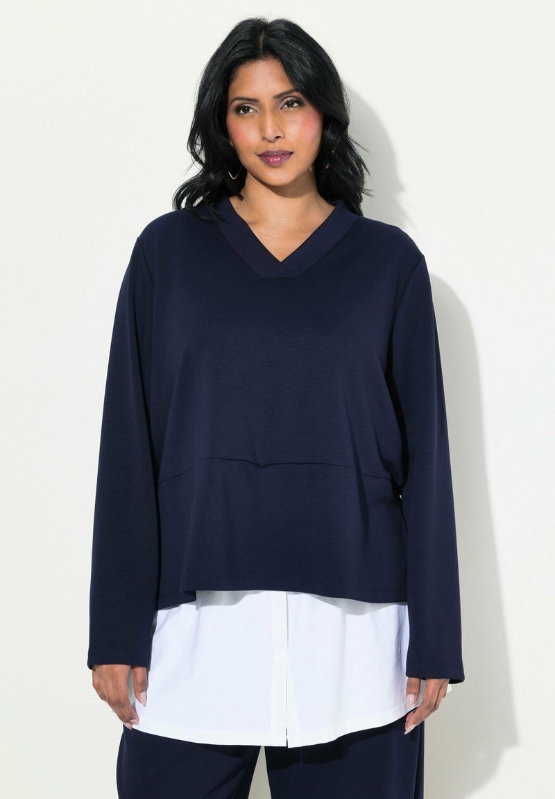 Camiseta de manga larga azul marino con escote en V, de corte relajado y apariencia de capas sobre una camisa blanca, con tejido suave y una ligera textura.