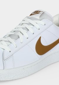 Λευκό δερμάτινο αθλητικό παπούτσι Nike με καφέ λογότυπο swoosh, λευκά κορδόνια και υφασμένο λευκό πέλμα, εμφανίζεται σε κοντινή γωνιακή όψη.