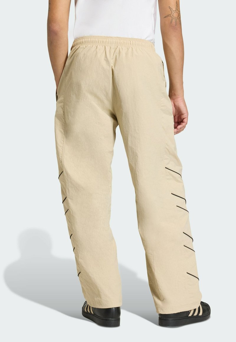Pantaloni beige con vita elasticizzata presentano spacchi laterali con dettagli neri, evidenziando una vestibilità ampia e un tessuto leggermente strutturato. Scarpe nere visibili.