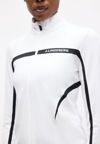 Γυναίκα που φοράει λευκό μπουφάν με φερμουάρ και μαύρες ρίγες, με το λογότυπο "J.LINDEBERG" στο στήθος και στα μανίκια, σε λευκό φόντο.