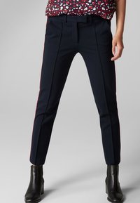 Pantalon taillé bleu marine avec des rayures latérales rouges contrastantes, présentant un devant plat, des passants de ceinture et un design ajusté. Tissu lisse et forme structurée.