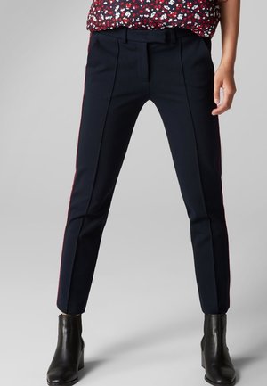 Pantalon taillé bleu marine avec des rayures latérales rouges contrastantes, présentant un devant plat, des passants de ceinture et un design ajusté. Tissu lisse et forme structurée.