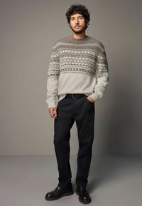 Pull léger gris avec motif géométrique en gris foncé, associé à un pantalon noir et des chaussures noires. Le pull présente des poignets côtelés.