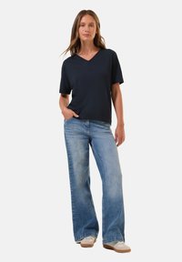 Námořnické tričko s výstřihem do V a krátkým rukávem, spárované s lehkými modrými širokými džínami. Modelka stojí s jednou rukou v kapse a má na sobě světlou obuv.