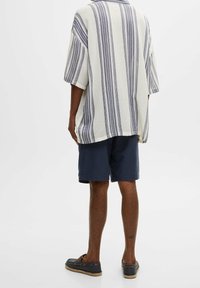 Camicia leggera a righe verticali blu e bianche, maniche corte e vestibilità ampia, abbinata a pantaloni corti navy e scarpe scure.