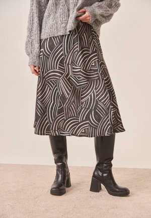 Mujer vestida con una falda midi estampada, suéter gris de punto grueso y botas negras de tacón hasta la rodilla, de pie sobre una alfombra beige.