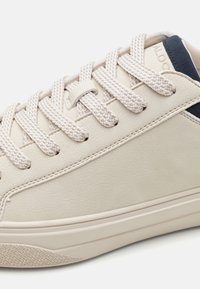 Beige sneaker with textured leather upper, round toe, and navy accent on heel. Features flat, woven laces and a subtle patterned outsole.