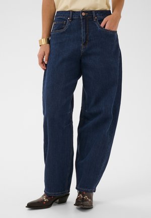 Persona che indossa jeans blu scuro larghi, camicia chiara, stivaletti neri ricamati con motivi floreali e un ampio braccialetto d'oro al polso sinistro.