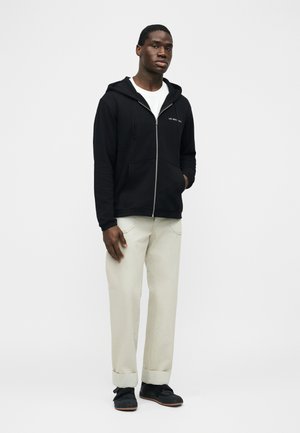 Nuori mies pukeutuneena mustaan vetoketjulliseen huppariin, valkoiseen t-paitaan, vaaleanbeigeihin käännettyihin housuihin ja mustiin tennareihin, seisoo yksinkertaisen vaalean taustan edessä.