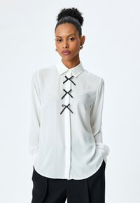 Blusa blanca de manga larga con cuello, que presenta tres detalles de cinta negra en el centro. Fabricada con un material ligero y suave.