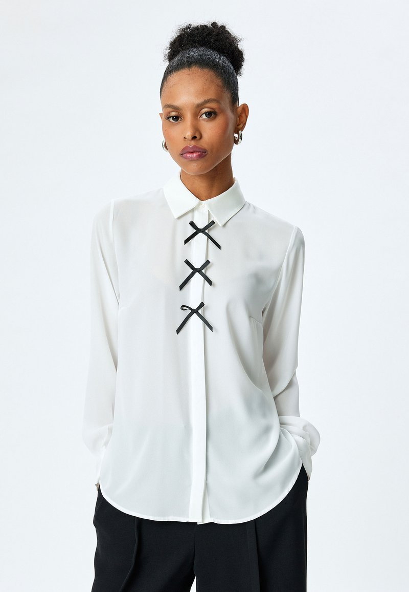 Blusa blanca de manga larga con cuello, que presenta tres detalles de cinta negra en el centro. Fabricada con un material ligero y suave.