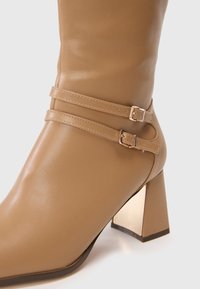 Bottine en cuir tan avec deux sangles horizontales et boucles. Présente un talon carré en métal et une surface lisse et polie.