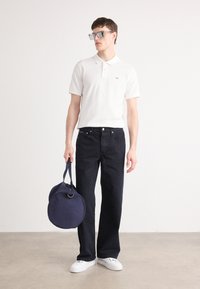 Modell som har på seg en hvit polo skjorte, mørk jeans og hvite joggesko, holder en marineblå rund duffelbag med topphåndtak og glidelås.