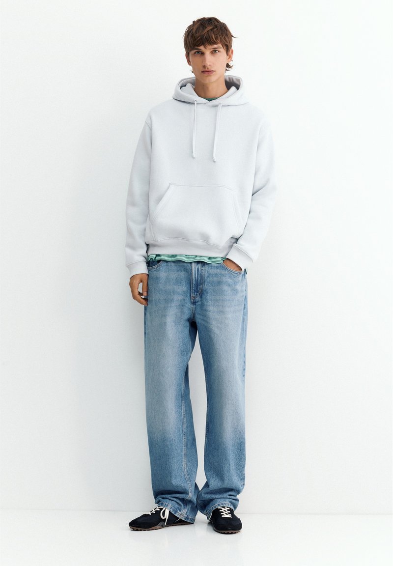 Jeune personne debout portant un sweat à capuche gris clair, un jean bleu ample et des baskets noires sur un fond blanc uni.