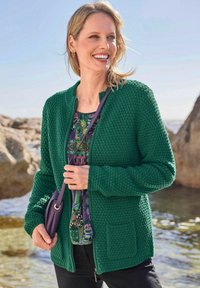 Grüner strukturierter Reißverschluss-Cardigan mit rundem Ausschnitt, kombiniert mit einem gemusterten Oberteil. Verfügt über eine Tasche auf der Vorderseite und hat ein glattes Finish.