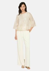 Top trasparente beige con motivo floreale e maniche larghe, abbinato a pantaloni bianchi a gamba larga e sandali metallici con cinturini.