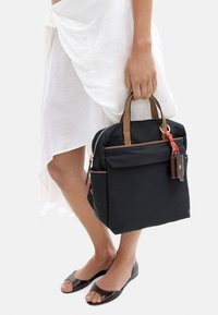 Czarny plecak materiałowy z brązowymi skórzanymi uchwytami, zamek błyskawiczny, przednia kieszeń i czerwona zawieszka w kształcie klucza, trzymany w ręce. Gładka tekstura.