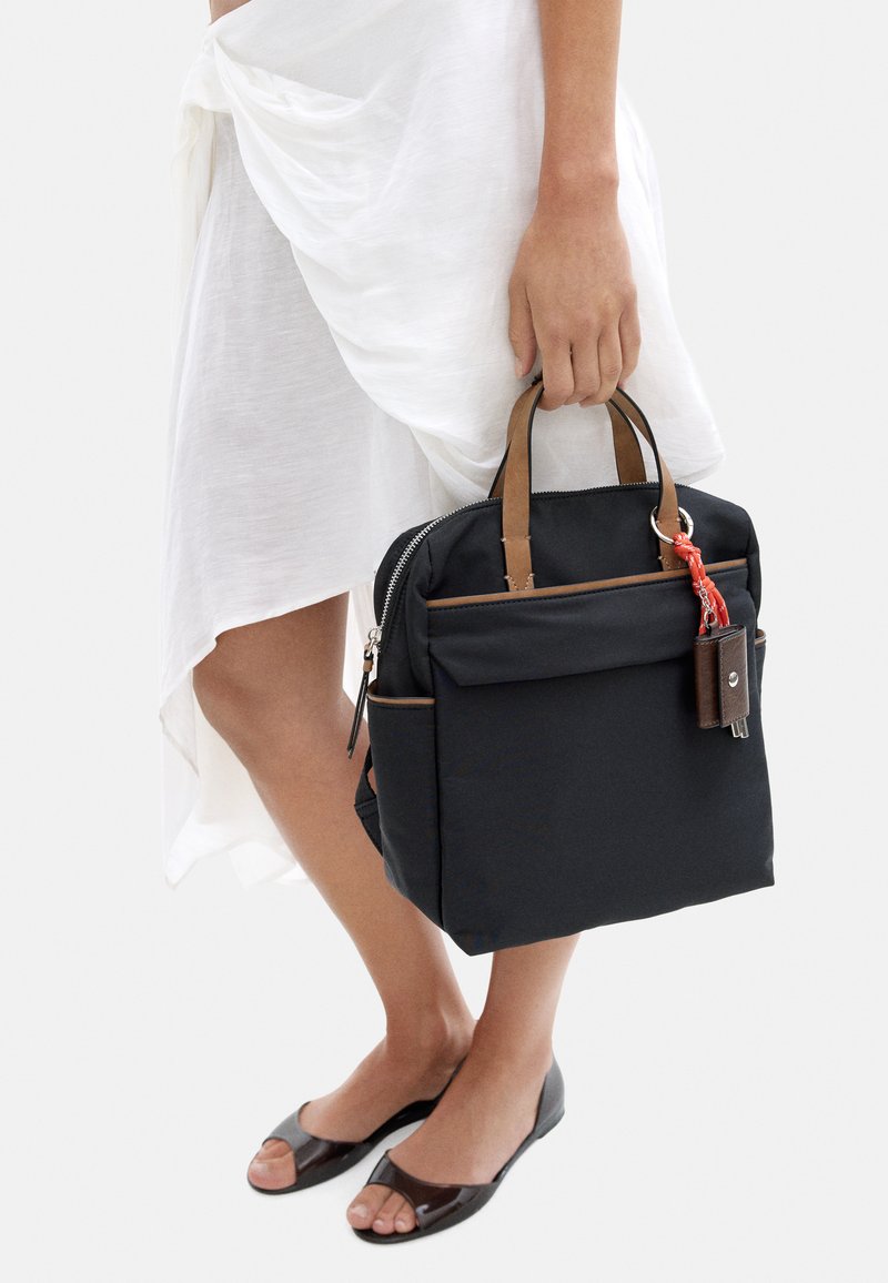 Czarny plecak materiałowy z brązowymi skórzanymi uchwytami, zamek błyskawiczny, przednia kieszeń i czerwona zawieszka w kształcie klucza, trzymany w ręce. Gładka tekstura.