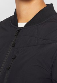 Svart vadderad bomberjacka med dragkedja, ribbad krage och slät tygtextur med minimala sömdetaljer.